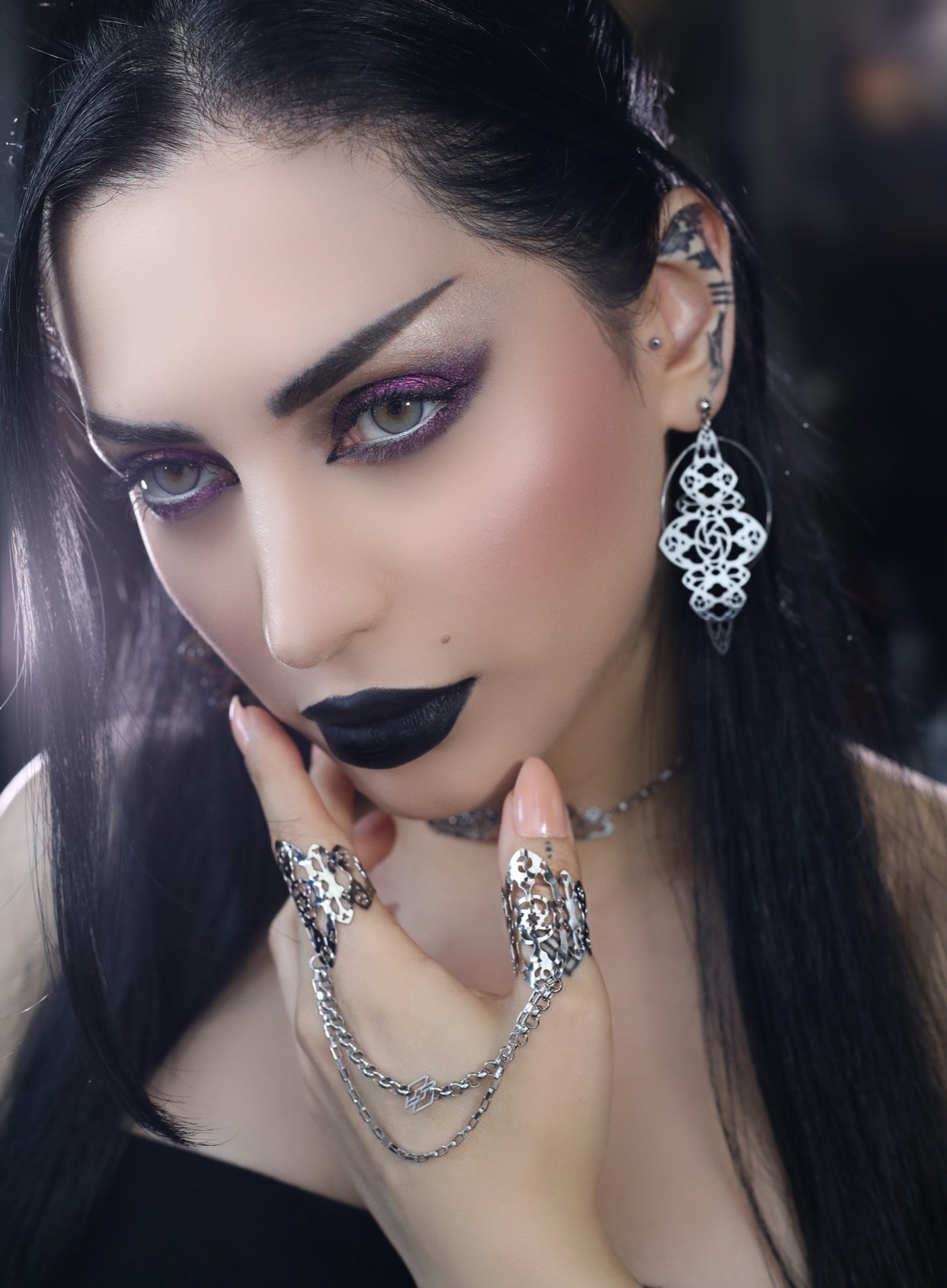 A striking gothic model with bold black lipstick and shimmering purple eye makeup showcases Myril Jewels' intricate accessories. She wears ornate white filigree earrings and matching rings connected by a delicate chain, exemplifying the dark-avantgarde aesthetic. Ideal for those embracing neo-gothic style or looking for bold witchcore accessories.