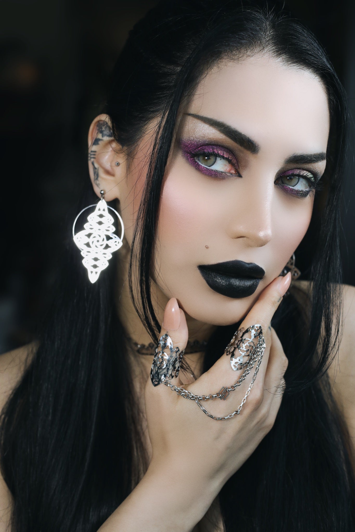 A striking gothic model with bold black lipstick and shimmering purple eye makeup showcases Myril Jewels' intricate accessories. She wears ornate white filigree earrings and matching rings connected by a delicate chain, exemplifying the dark-avantgarde aesthetic. Ideal for those embracing neo-gothic style or looking for bold witchcore accessories.