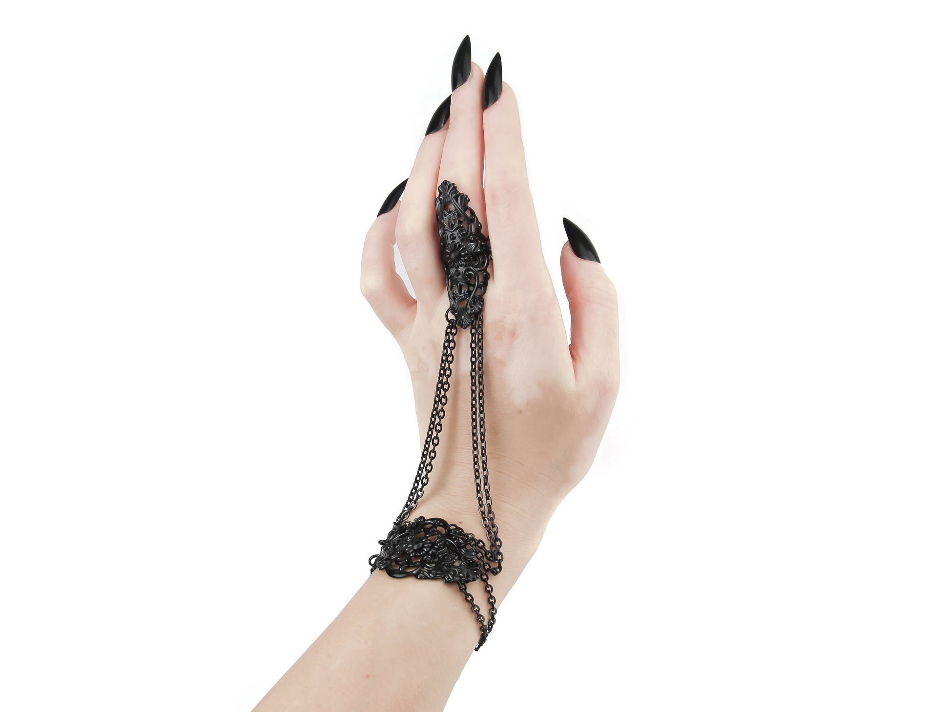 A sophisticated, handcrafted jewelry piece in elegant black is adorned with a gothic design. The back of the hand is covered in detailed filigree metalwork, with graceful chains connecting the bracelet part to the ring on the middle finger. The hand is shown with sharply pointed black manicured nails, adding to the dark allure of the piece. The white background accentuates its beauty and sophistication.