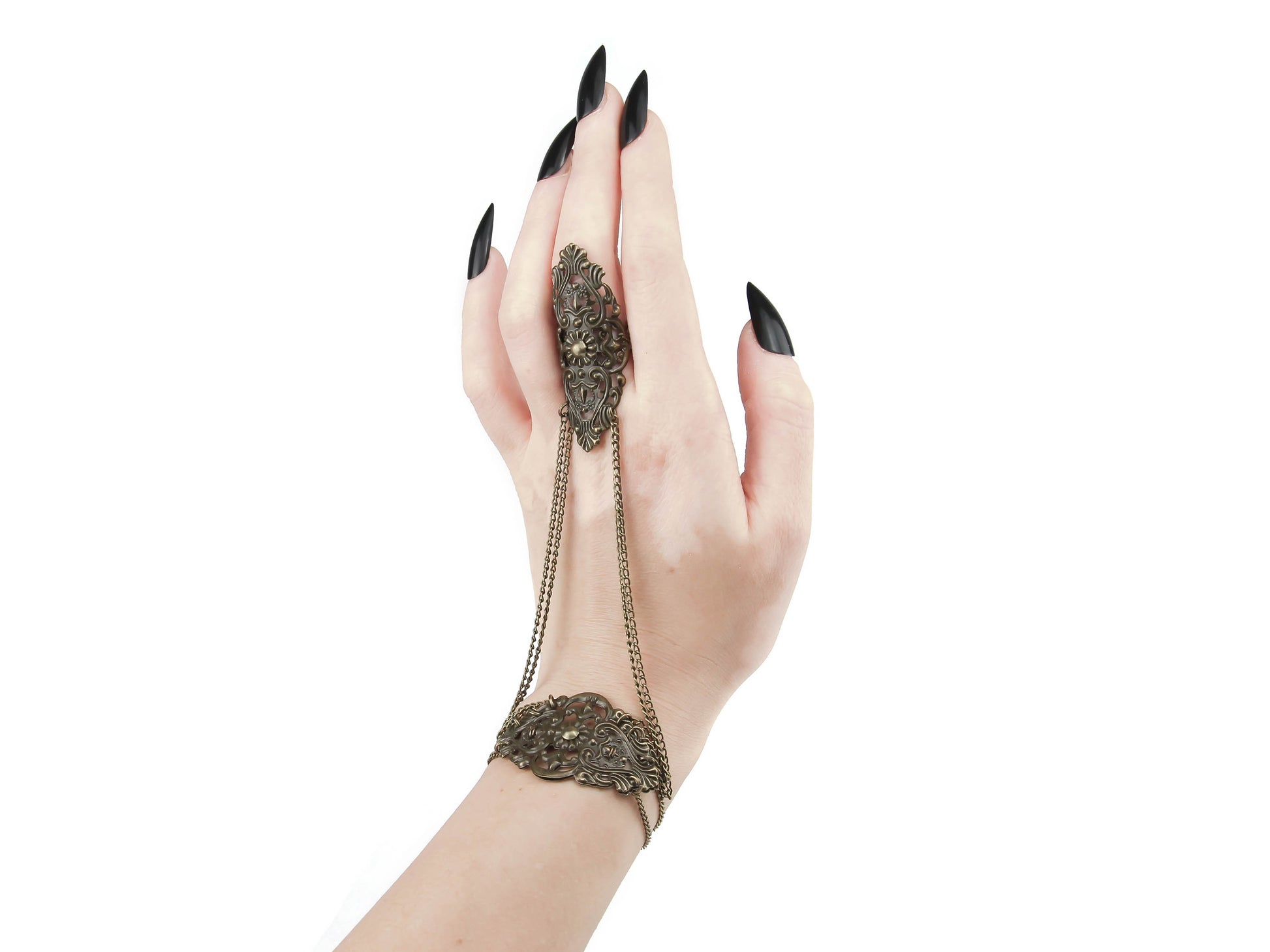 A stunning bronze hand chain adornment with a gothic-inspired design is shown off on a hand adorned with sharp, black manicured nails, capturing the essence of goth fashion. The piece boasts intricate filigree metalwork that adorns the back of the hand, while chains elegantly connect the bracelet portion to the middle finger ring. The background is a crisp white, emphasizing the refined and mysterious charm of this meticulously crafted creation.