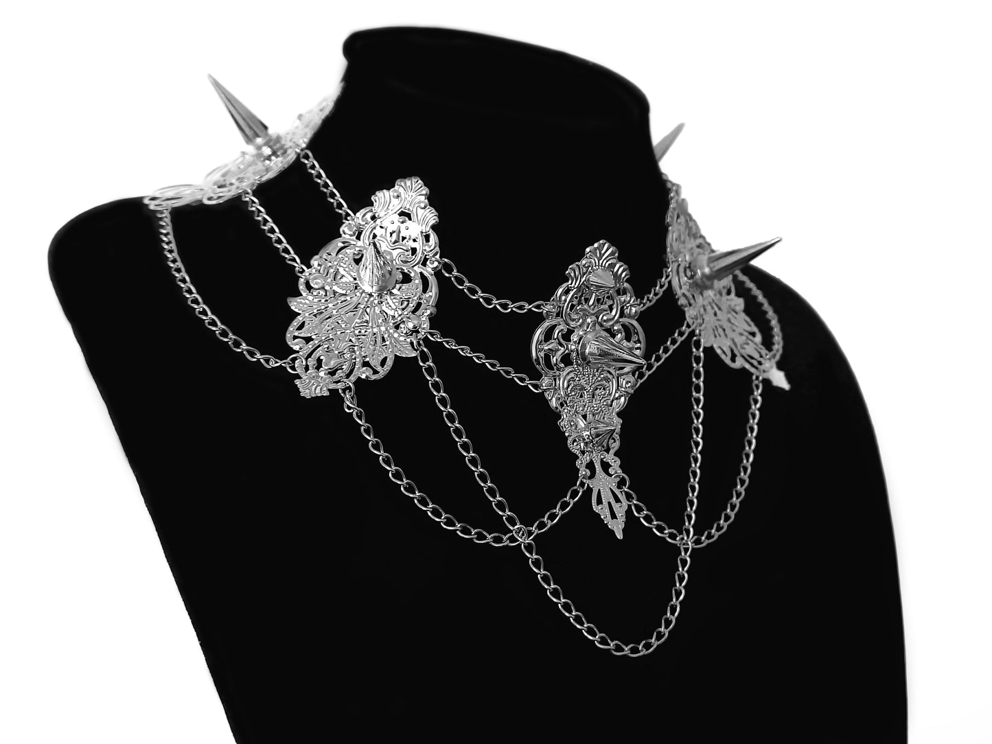 An intricate studded choker by Myril Jewels, featuring ornate silver filigree and sharp spikes, draped with delicate chains. This bold piece captures the essence of neo-gothic elegance, ideal for witchcore enthusiasts and a standout accessory for festival or rave attire.