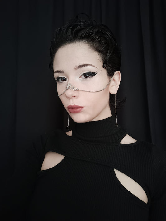 A poised model with a sleek updo showcases a delicate Myril Jewels silver nose chain against a dark backdrop. The chain's intricate design adds a touch of gothic-chic to the avant-garde aesthetic, perfect for anyone embracing a neo-goth, Witchcore, or whimsigoth lifestyle.