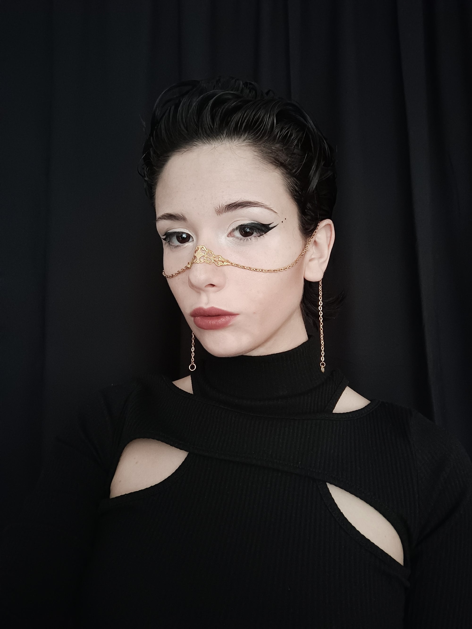 In a striking black and white portrait, a model from Myril Jewels exhibits a delicate gold nose chain against a dark backdrop. The accessory complements a neo-goth look, ideal for those who appreciate gothic-chic jewelry, making it a versatile choice for everyday wear, Halloween, or as a unique goth girlfriend gift.