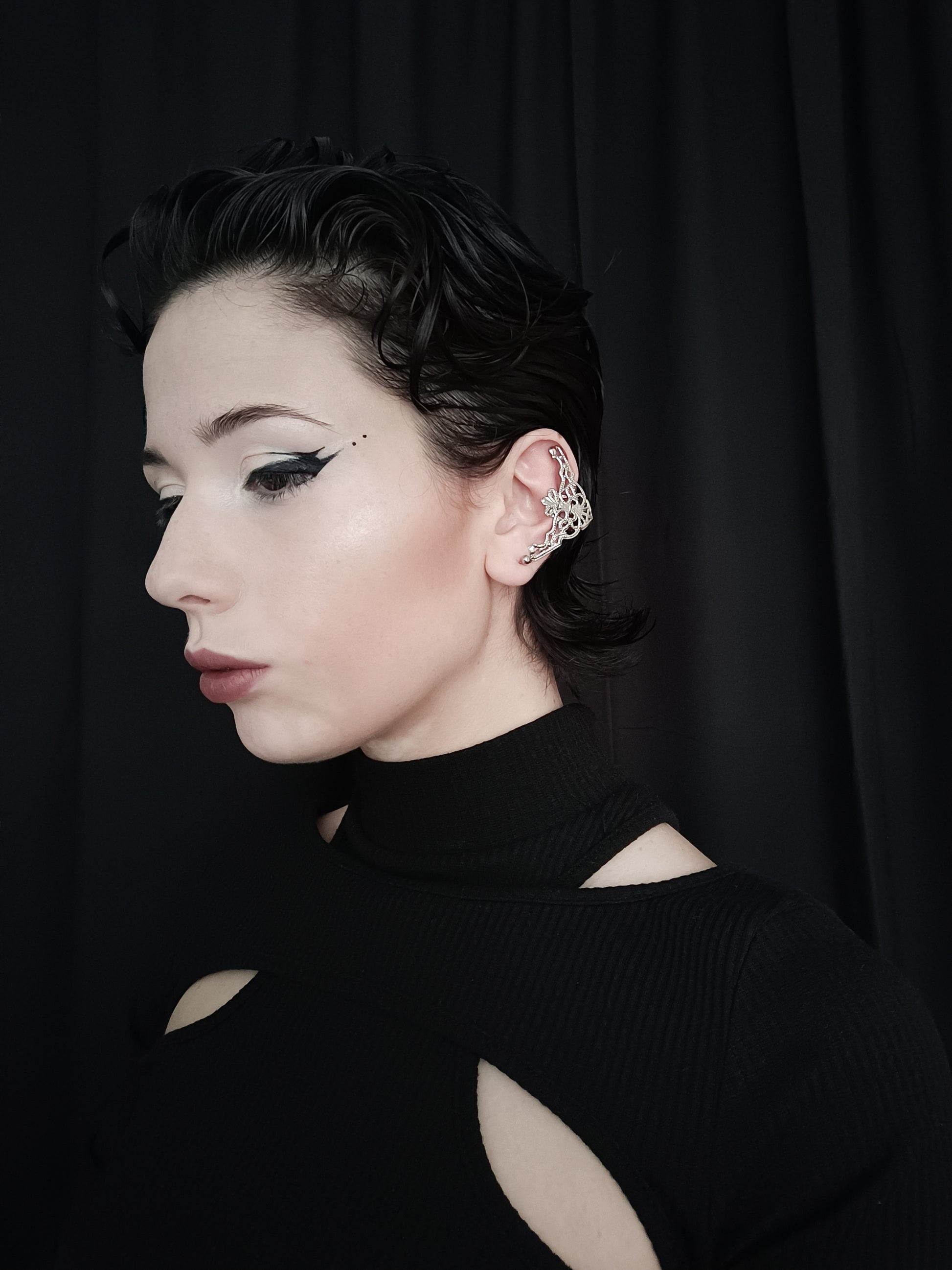 A profile view of a model wearing Myril Jewels cuff earrings, showcasing a neo-goth design with intricate filigree details. The earrings, resembling elegant gothic arches, complement the model's dark avant-garde style. This gothic-chic accessory is perfect for those embracing a whimsigoth or witchcore aesthetic, versatile enough for everyday wear, and striking for a Halloween or festival ensemble. The model's sleek hairstyle and bold eyeliner accentuate the earrings