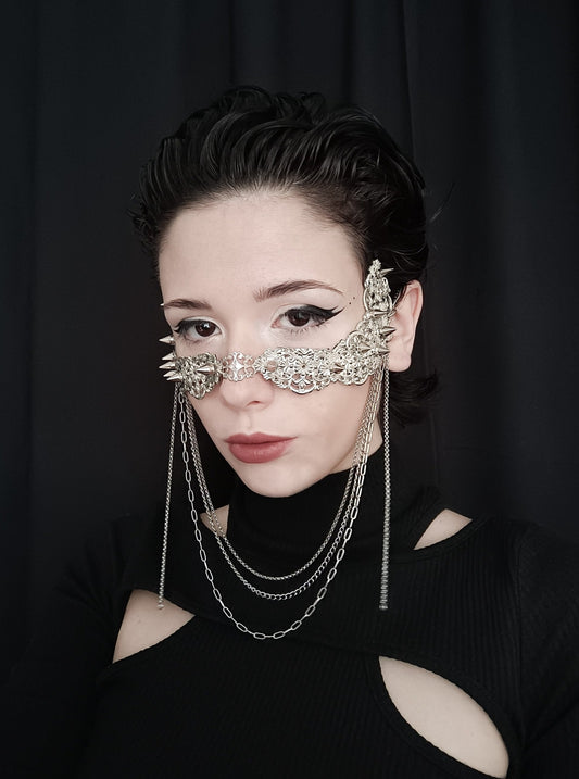 A model poses in a unique Myril Jewels nose mask, adorned with studs and cascading chains, evoking a neo-goth elegance. This striking piece blends dark-avantgarde with gothic-chic, perfect for the alternative style enthusiast. Ideal for adding an edgy touch to Halloween costumes, witchcore fashion, or as a statement accessory at a rave party or festival, it exemplifies Myril's commitment to innovative, handmade jewelry.