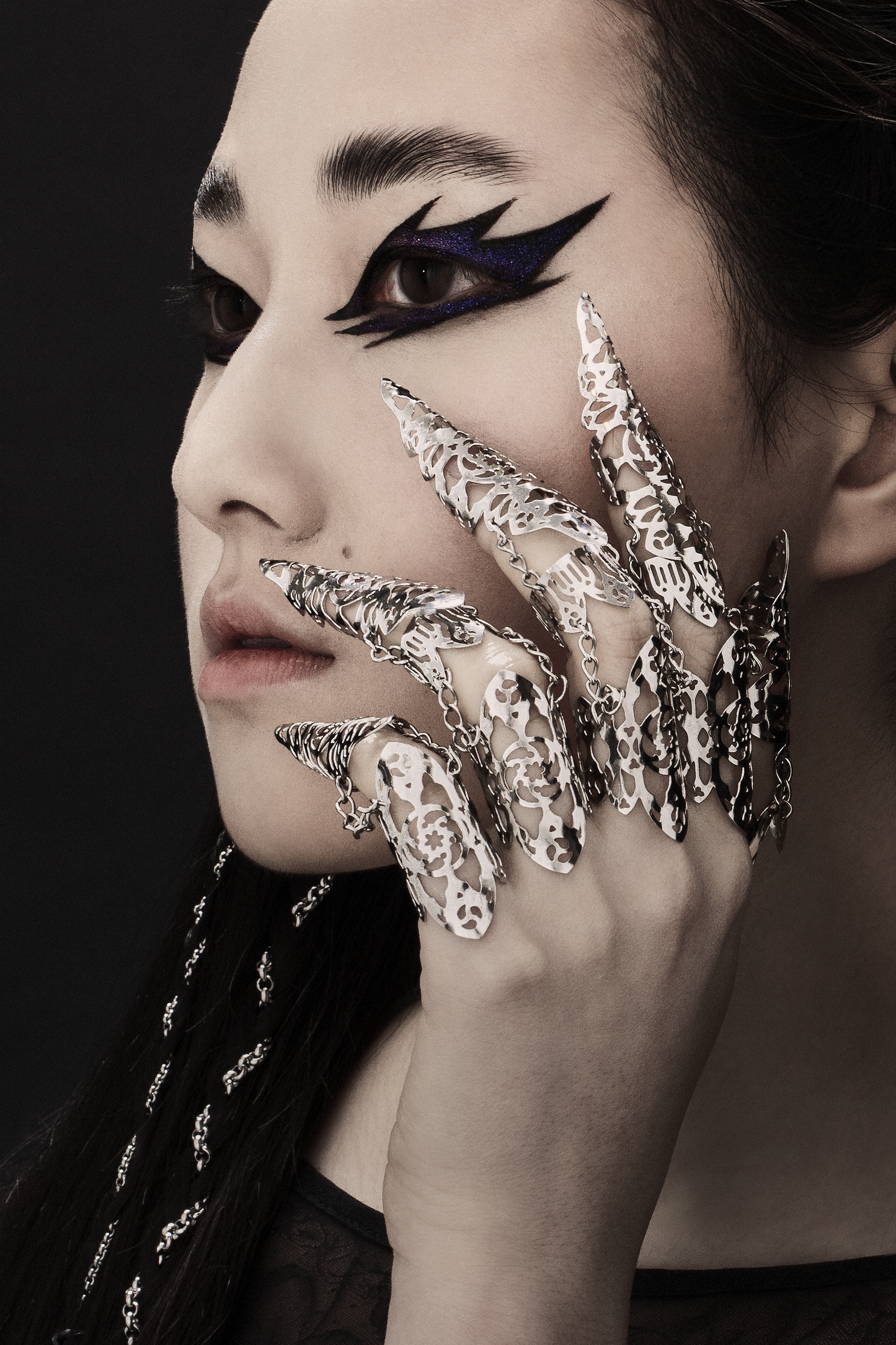 A portrait of an individual with dramatic black winged eyeliner and a full set of full-finger claw rings from Myril Jewels. The jewelry’s intricate patterns and chain links embody the brand’s dark, avant-garde neo-goth aesthetic. Perfect for gothic, whimsigoth, and witchcore enthusiasts, these claw rings make a bold statement at any Halloween or rave party and are a distinctive gift for lovers of gothic-chic and minimal goth styles.