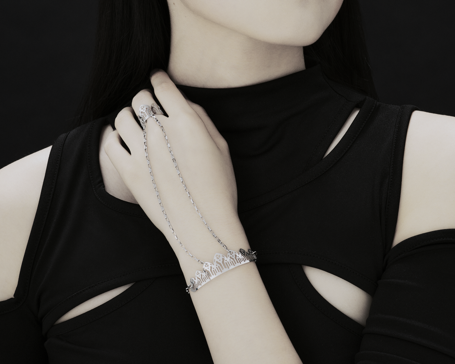 Discover Myril Jewels' exquisite craftsmanship with this hand chain bracelet ring, embodying a gothic allure. The image showcases an elegant silver filigree bracelet, seamlessly transitioning into a matching ring via a delicate chain. This sophisticated piece contrasts beautifully against the model's black sleeveless top, highlighting the meticulous cut-out design that resonates with the dark-avantgarde theme.
