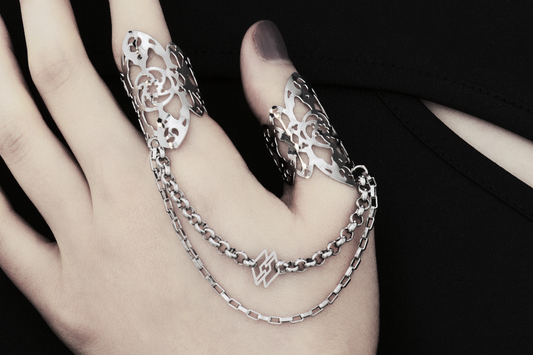 A hand models Myril Jewels' handcrafted ring featuring elaborate cut-out designs that echo a gothic architectural aesthetic, suited for lovers of dark-avantgarde fashion. The rings are interconnected with sleek chains, creating a bold, edgy look. This collection exemplifies the Italian craftsmanship and innovative style synonymous with the Myril Jewels brand, catering to the tastes of the alternative and gothic jewelry community.