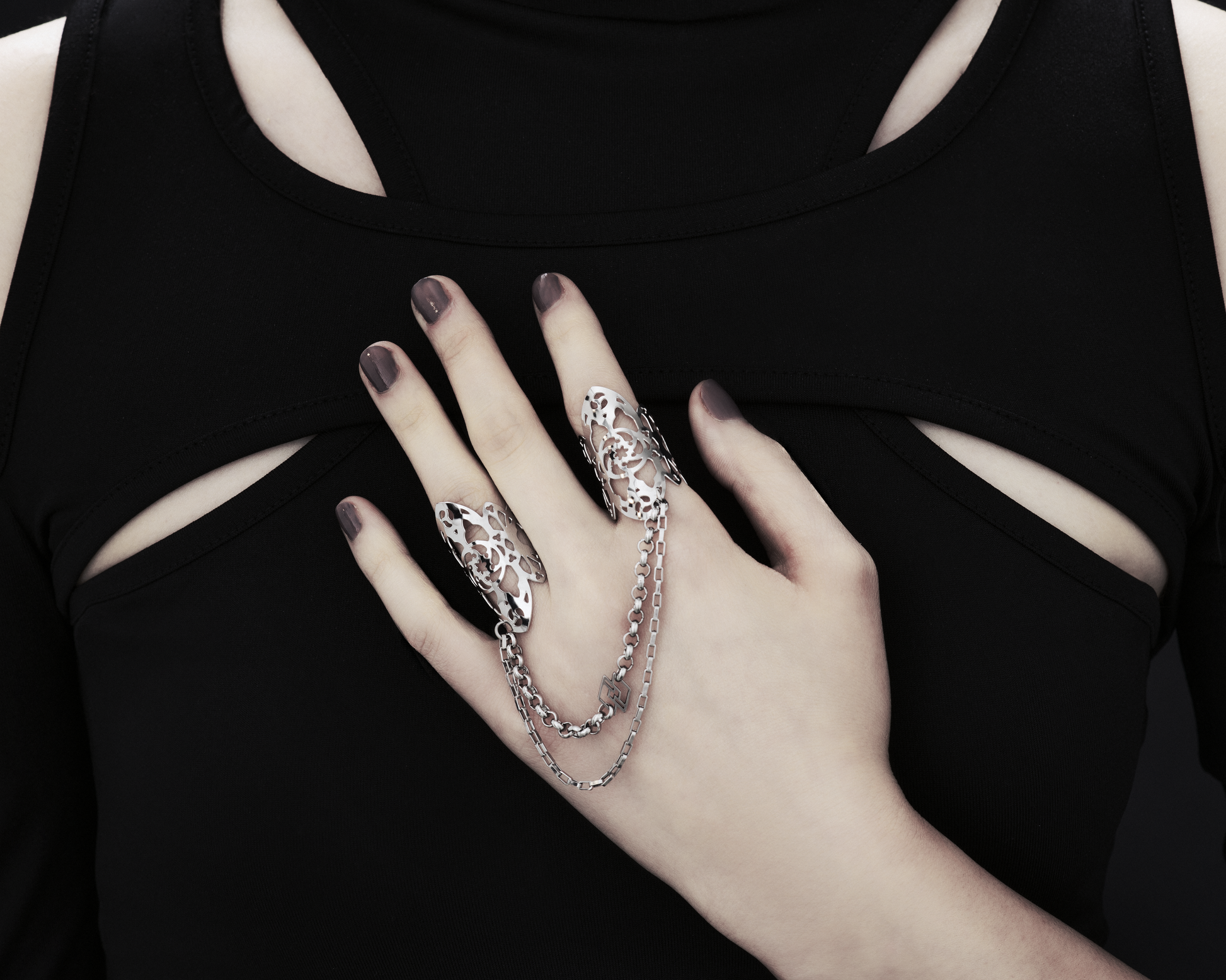 A hand models Myril Jewels' handcrafted ring featuring elaborate cut-out designs that echo a gothic architectural aesthetic, suited for lovers of dark-avantgarde fashion. The rings are interconnected with sleek chains, creating a bold, edgy look. This collection exemplifies the Italian craftsmanship and innovative style synonymous with the Myril Jewels brand, catering to the tastes of the alternative and gothic jewelry community.