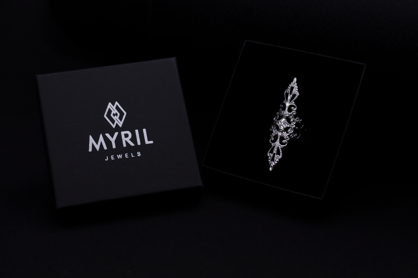 A sleek Myril Jewels branded box open on a dark surface, revealing a sophisticated silver filigree ring inside, showcasing the brand's refined gothic jewelry collection.