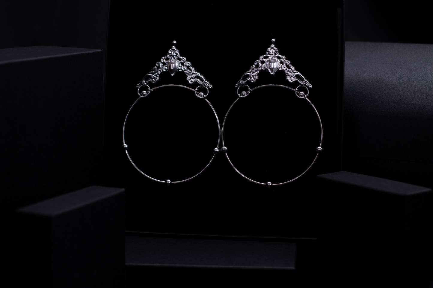 A pair of Myril Jewels' bold gothic hoop earrings rest against a plush black backdrop, exuding an air of dark sophistication. These wide hoops are adorned with intricate silver filigree and a central gemstone, reflecting the brand's commitment to avant-garde style. Ideal for the goth girlfriend, they're perfect for festivals, everyday wear, or as a statement piece for drag queens.