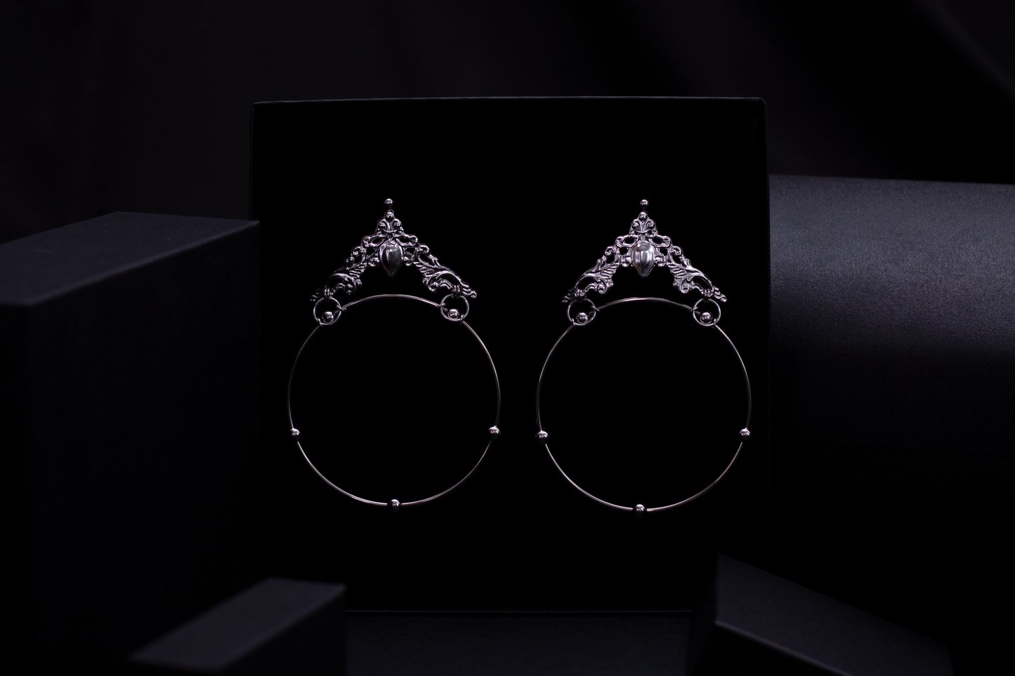 A stunning display of Myril Jewels' craftsmanship, this image showcases bold and wide gothic hoop earrings with ornate detailing. The earrings are set against a dark backdrop, highlighting their intricate design and the shimmering central gemstone. Ideal for those who covet neo-gothic style, these earrings are perfect as a statement piece for Halloween, goth girlfriend gifts, or as a distinctive accessory for any alternative wardrobe.