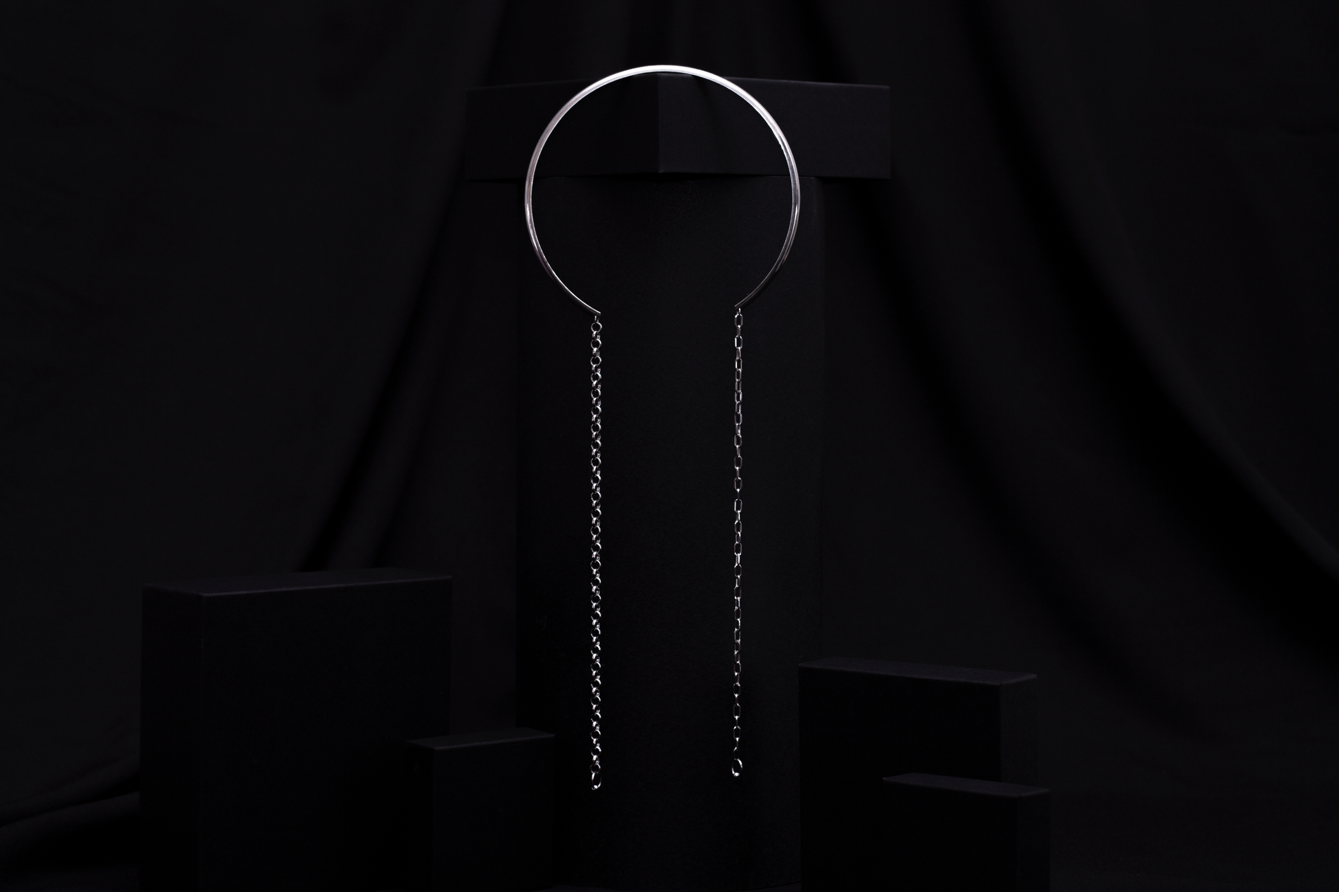 Displayed against a black backdrop, this Myril Jewels piece features a rigid, minimal necklace with long hanging chains, encapsulating the essence of minimal goth. This refined item is ideal for adding an avant-garde touch to any outfit, suitable for everyday wear or as a distinctive accessory for special events like festivals.