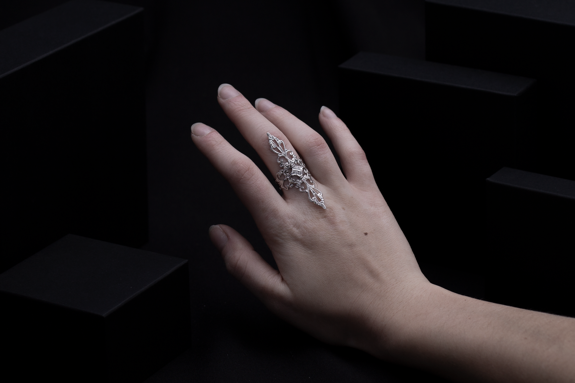 A woman's hand elegantly displaying a silver filigree full-finger ring against a dark background with geometric shapes, embodying a sophisticated gothic style.