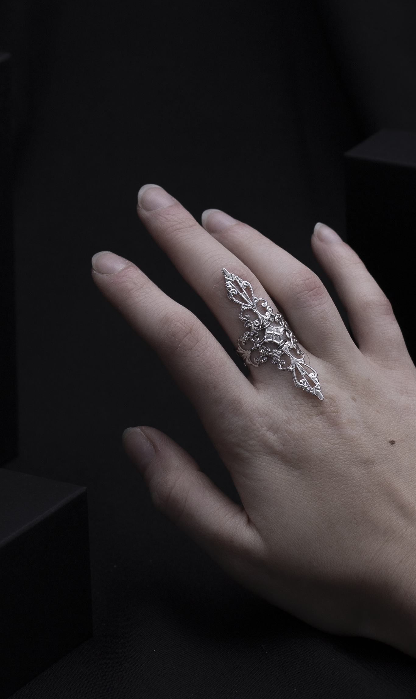 A woman's hand elegantly displaying a silver filigree full-finger ring against a dark background with geometric shapes, embodying a sophisticated gothic style.