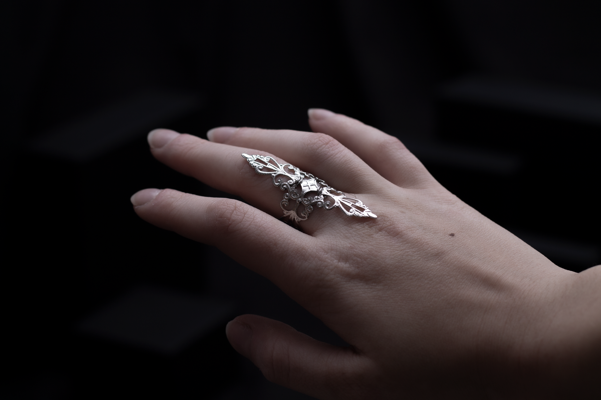 A woman's hand elegantly displaying a silver filigree full-finger midi ring against a dark background with geometric shapes, embodying a sophisticated gothic style.