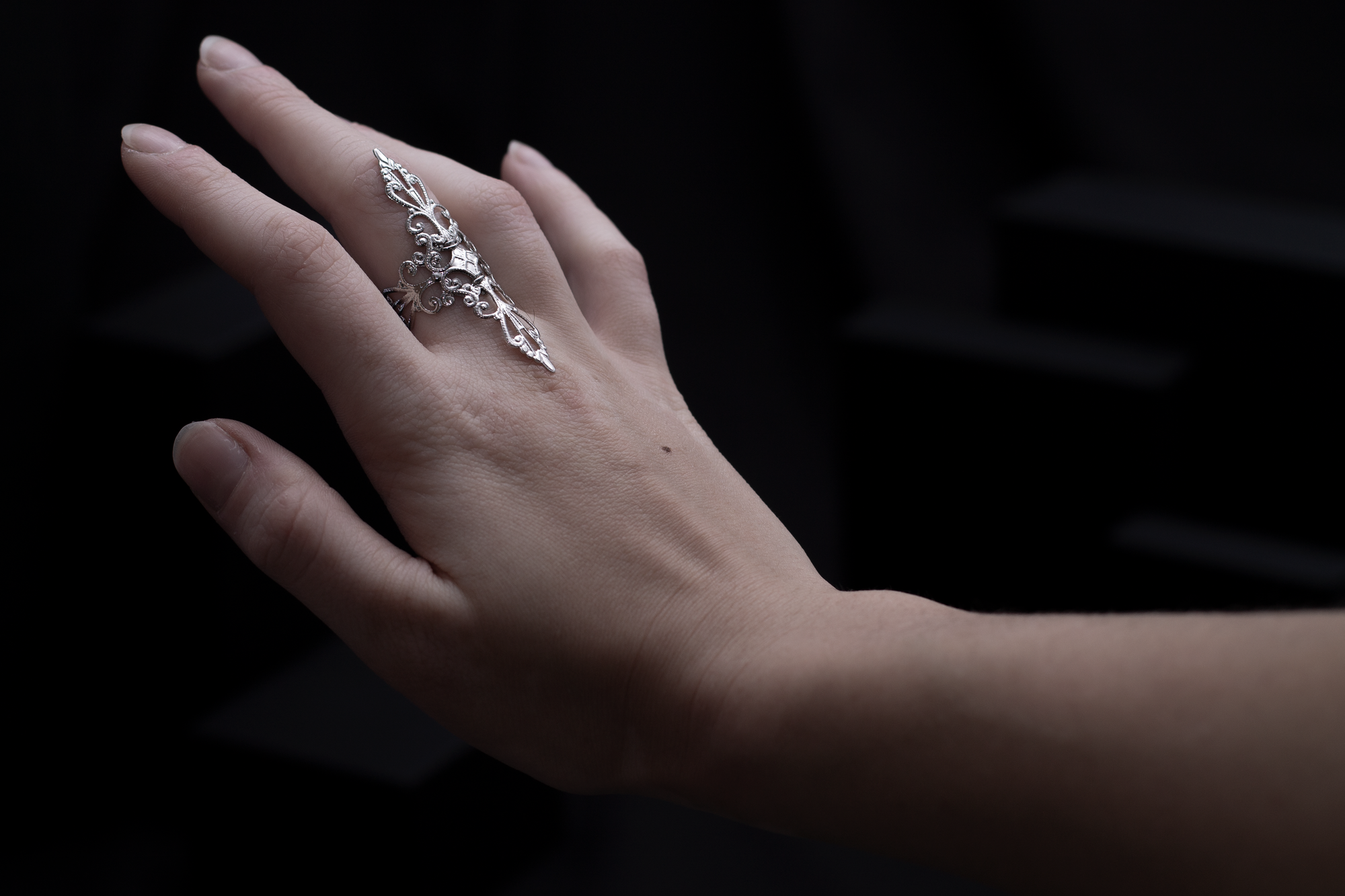 A woman's hand elegantly displaying a silver filigree full-finger ring against a dark background with geometric shapes, embodying a sophisticated gothic style.