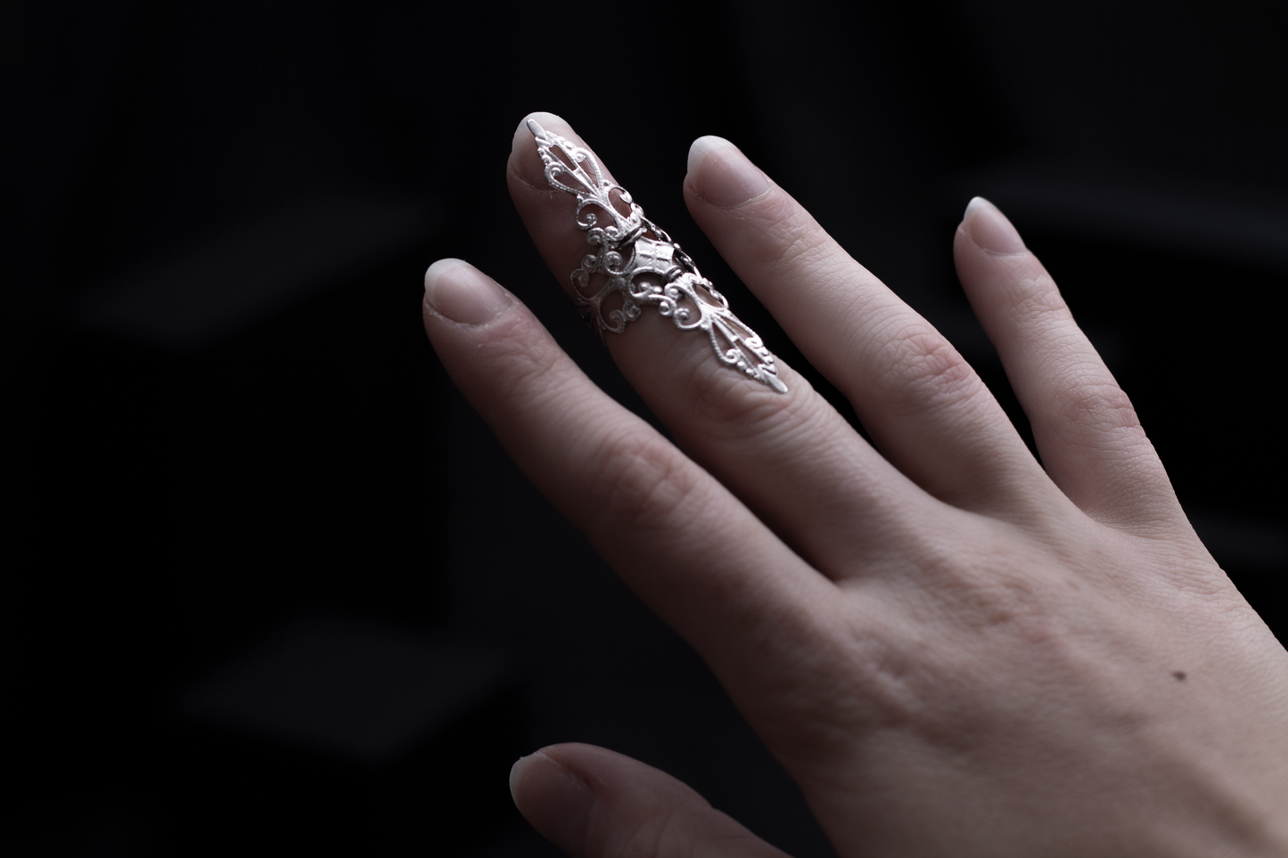 A woman's hand elegantly displaying a silver filigree full-finger midi ring against a dark background with geometric shapes, embodying a sophisticated gothic style.