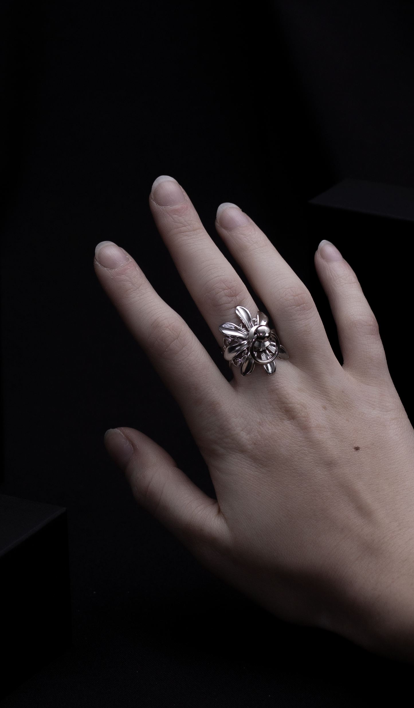 A solitary hand displays a Myril Jewels flower ring with o-ring stud in the middle, a masterpiece of dark-avantgarde jewelry with a punk twist. The ring's silver sheen and intricate floral design embody the luxurious, gothic aesthetic, a signature of Myril Jewels, catering to those with a penchant for bold, alternative fashion.