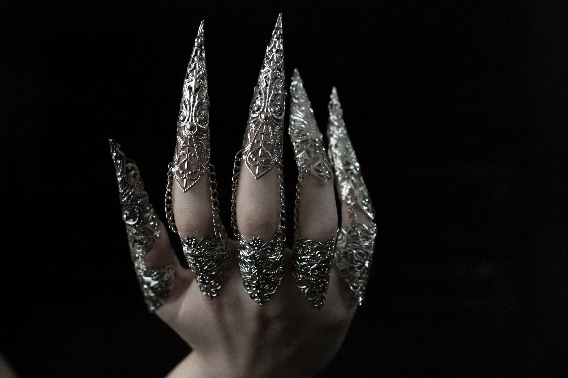 A hand is strikingly adorned with a full set of Myril Jewels rings, each extending into long, ornate claws that exemplify a dramatic neo-goth style. Crafted with intricate silver detailing, these rings are perfect for dark-avantgarde enthusiasts and make a bold statement piece for Halloween, punk events, and drag queen performances. They also complement a gothic-chic, whimsigoth, or witchcore aesthetic, suitable for everyday wear, and are a dazzling addition to rave party and festival jewelry collections.