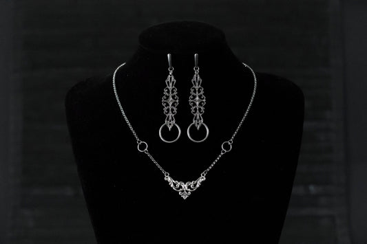 A Myril Jewels gothic necklace and matching earrings set is elegantly displayed on a black velvet mannequin. The silver necklace features a delicate filigree design, culminating in a classic gothic arch motif, paired with earrings that echo this design with an added hoop. Perfect for those who appreciate neo-goth jewelry, this set adds a touch of gothic-chic to any attire, making it ideal for Halloween, witchcore expressions, or as sophisticated everyday wear for the avant-garde individual.