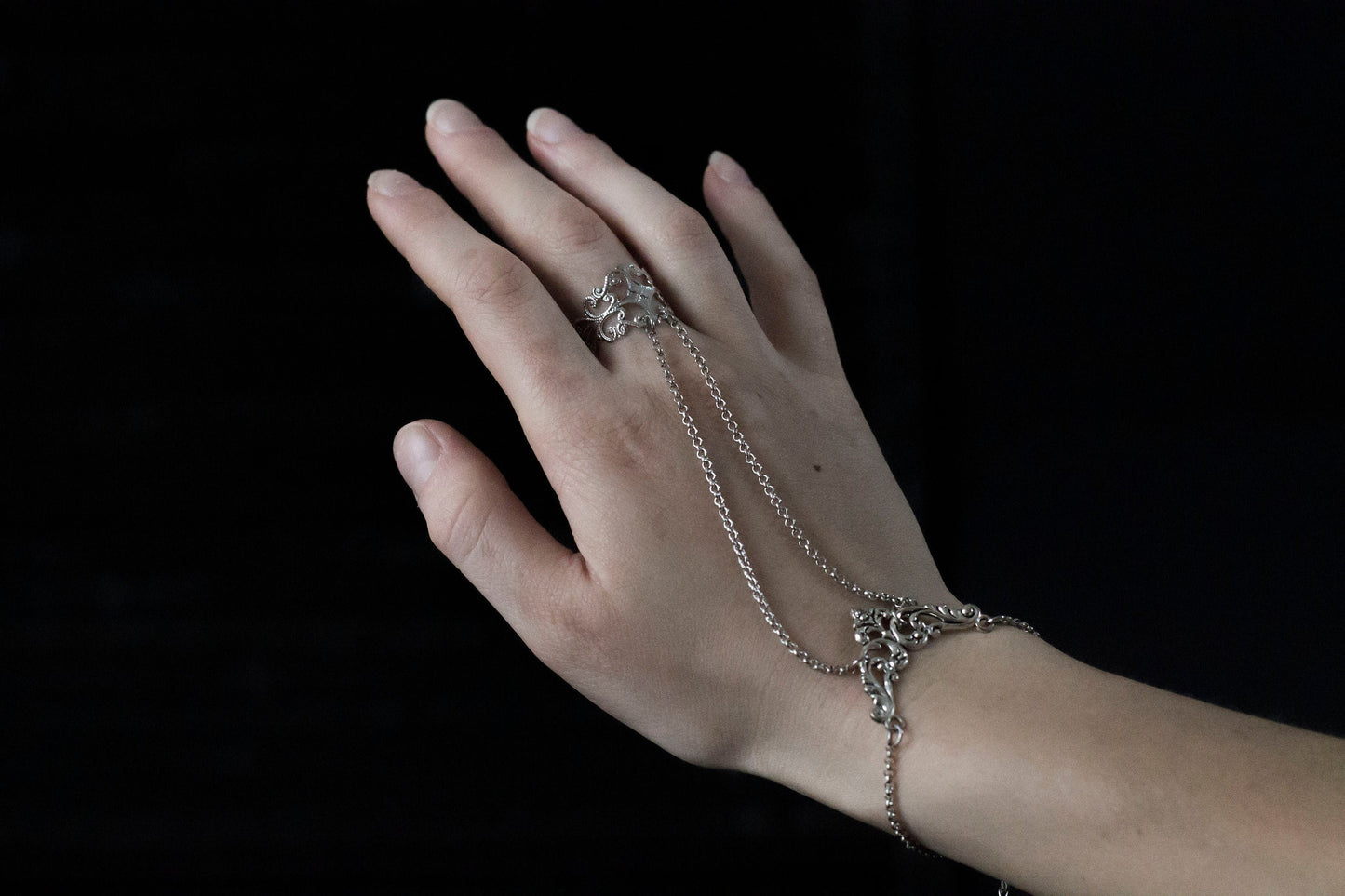 Elegant Myril Jewels hand chain bracelet ring displayed on a slender hand, portraying dark avant-garde luxury. The intricate silver filigree design, inspired by Neo Gothic floral style, cascades from a delicate ring down to a matching wrist cuff, creating a sophisticated, gothic-chic look. This jewelry is perfect for Halloween, everyday wear for the minimalist goth, or as an eye-catching accessory for rave parties and festivals.