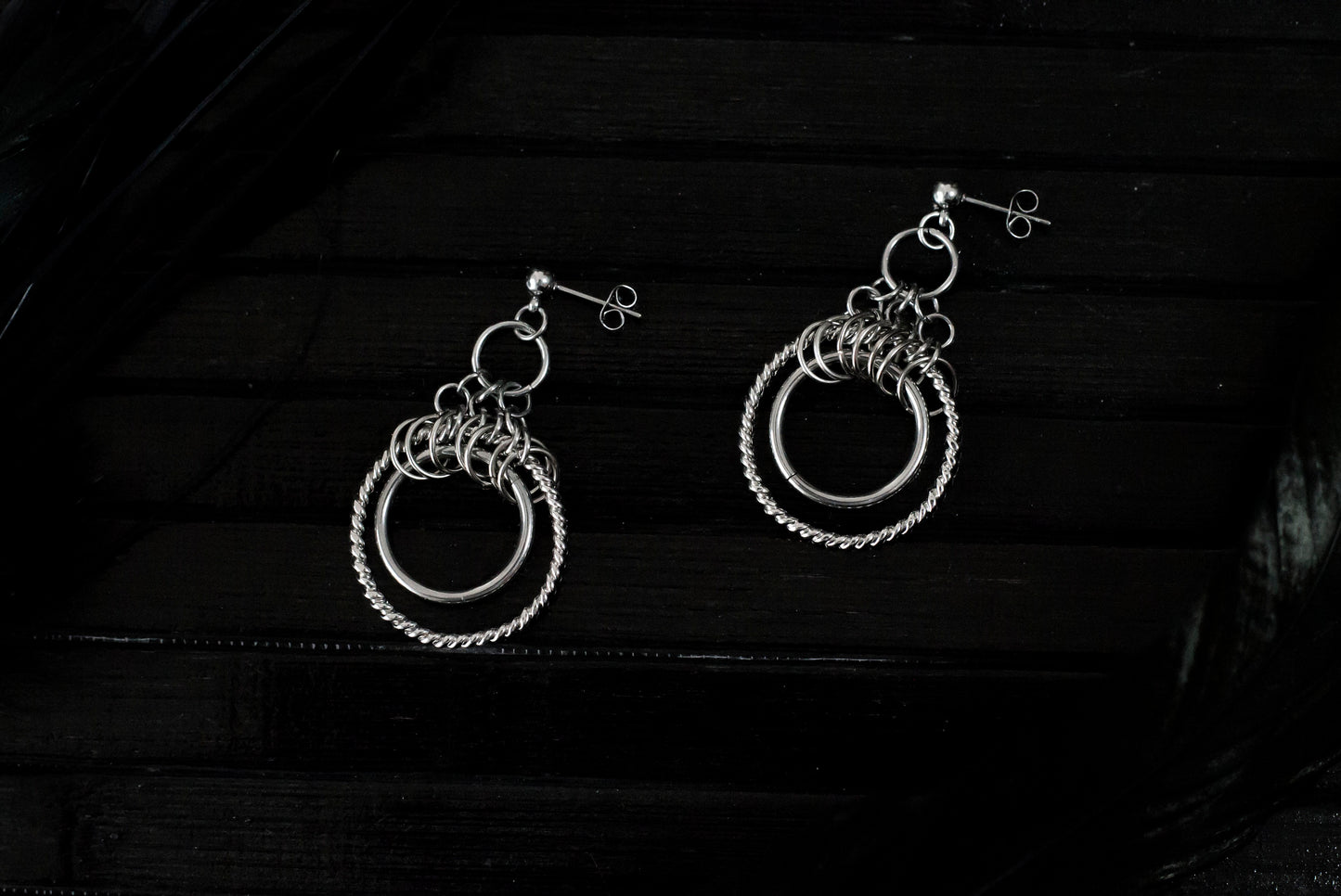 The image displays a pair of double hoop earrings with a sophisticated design. The earrings feature two concentric circles; the inner hoop is smooth and polished, while the outer hoop has a twisted rope-like texture, enhancing its visual appeal. Above the hoops, there is a cluster of smaller rings adding complexity to the design. These earrings, likely from Myril Jewels, reflect a gothic-chic style that merges elegance with an edgy touch, making them suitable for various occasions