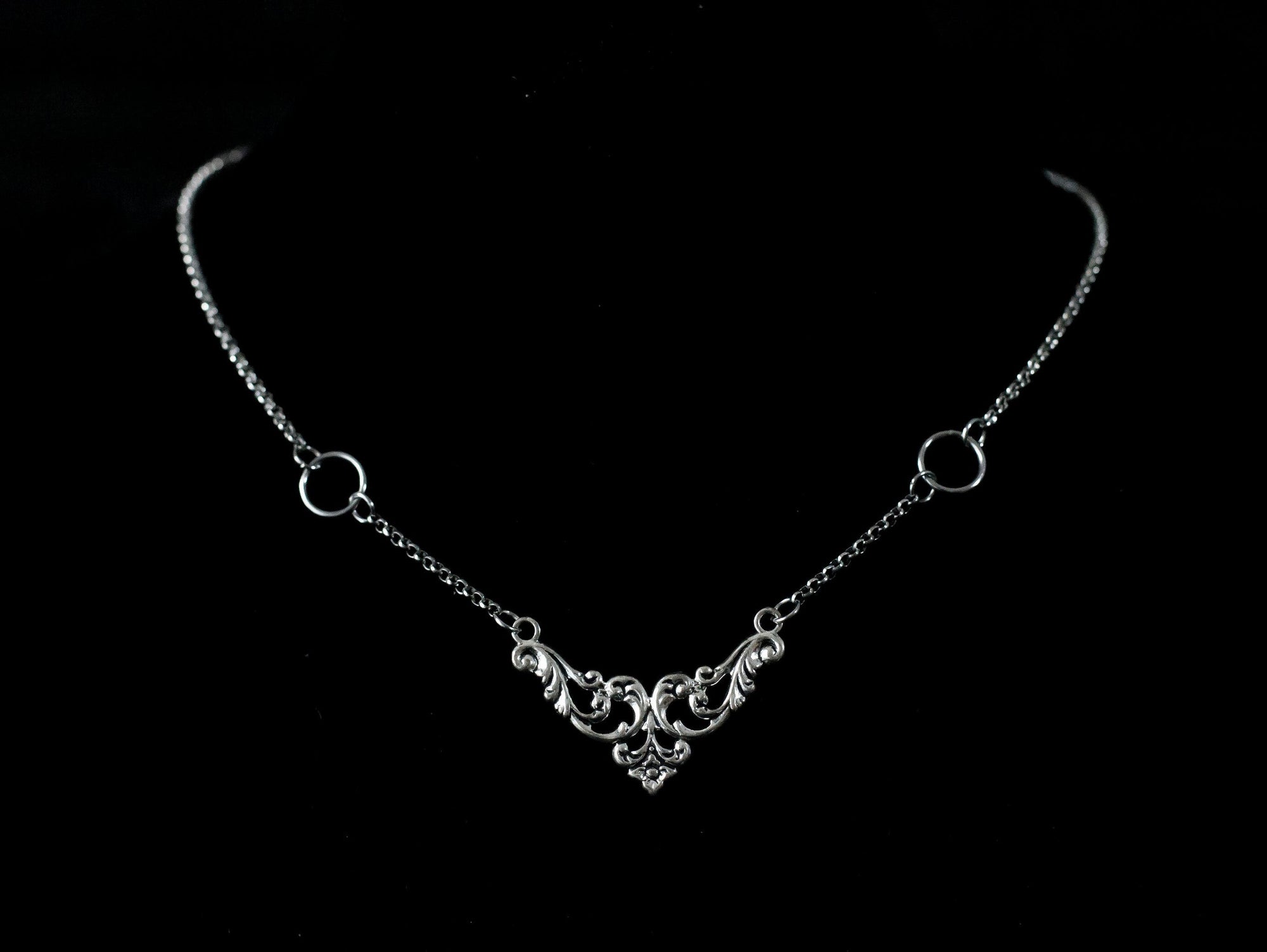 Displayed on a black velvet mannequin is a Myril Jewels elegant gothic necklace, capturing the essence of neo-goth fashion. The necklace, with a fine silver chain, features ornate filigree detailing and symmetrical O-ring accents, embodying the dark-avantgarde aesthetic. This piece is designed for gothic and alternative style aficionados, seamlessly blending with Halloween, punk, and gothic-chic ensembles, and is a sophisticated choice for whimsigoth, witchcore, or minimal goth everyday wear