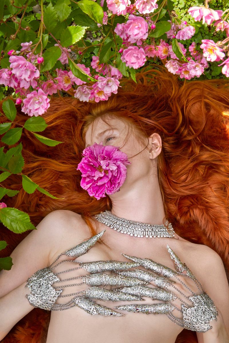 In a vibrant garden, a model with fiery red hair becomes one with nature, her face obscured by a deep pink bloom, embodying the whimsical goth aesthetic. Adorned in Myril Jewels' intricate silver metal glove with claw rings, the ensemble radiates neo-gothic elegance, perfect for gothic-chic or witchcore-inspired looks, and makes a statement piece for Halloween or punk fashion events.