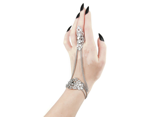An elegant silver hand chain jewelry piece with a gothic design is displayed on a hand with sharply pointed, black manicured nails, typical of a goth aesthetic. The jewelry features elaborate and detailed filigree metalwork that covers the back of the hand, with chains gracefully draping between the bracelet part and the ring on the middle finger. The background is pure white, highlighting the sophistication and dark allure of the handcrafted piece