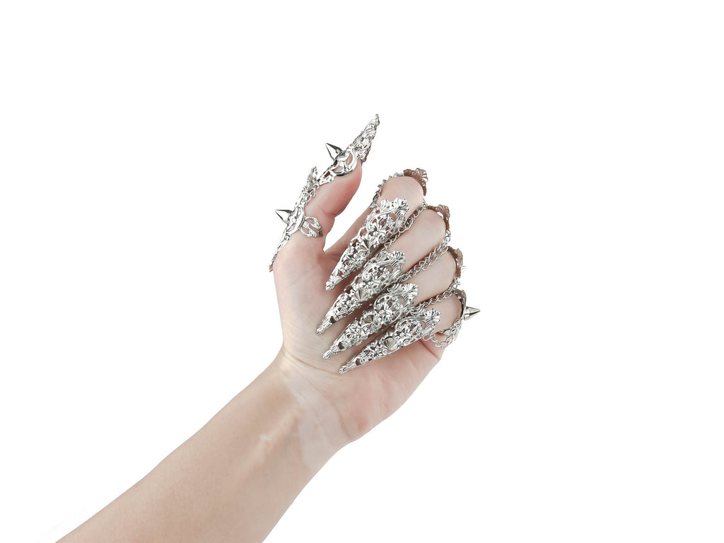  A hand is fully adorned with Myril Jewels' studded nail claw rings, each featuring a detailed silver design with sharp, edgy protrusions. These rings showcase a bold neo-goth aesthetic, perfect for dark-avantgarde fashion enthusiasts. The set is an ideal choice for creating a dramatic look at Halloween events, embodying the gothic-chic and whimsigoth spirit, and is also suited for witchcore, everyday wear, or as statement jewelry for rave parties and festivals.