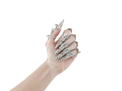  A hand is fully adorned with Myril Jewels' studded nail claw rings, each featuring a detailed silver design with sharp, edgy protrusions. These rings showcase a bold neo-goth aesthetic, perfect for dark-avantgarde fashion enthusiasts. The set is an ideal choice for creating a dramatic look at Halloween events, embodying the gothic-chic and whimsigoth spirit, and is also suited for witchcore, everyday wear, or as statement jewelry for rave parties and festivals.
