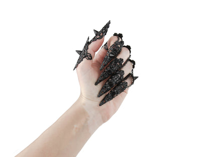 A hand is dramatically extended, displaying a full set of Myril Jewels studded nail claw rings in black. Each claw ring is elaborately designed with spikes and filigree, reflecting a dark-avantgarde and neo-goth aesthetic. These bold pieces are tailored for gothic, punk, and alternative style enthusiasts, perfect for Halloween, and suitable as an expressive accessory for everyday wear. They embody the gothic-chic look, enhancing whimsigoth and witchcore outfits, and making a statement at rave parties