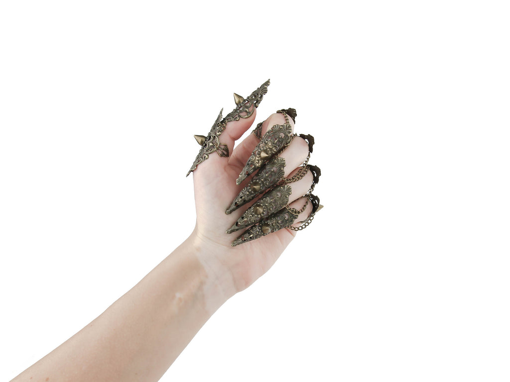A hand is dramatically adorned with a full set of Myril Jewels studded nail claw rings in bronze. The claw rings feature elaborate filigree with pointed studs, invoking a bold neo-goth aesthetic. These edgy, dark-avantgarde pieces are perfect for those who embrace gothic and alternative styles, making them an ideal choice for Halloween, punk-inspired outfits, or as a statement addition to a whimsigoth or witchcore look. They also serve as eye-catching accessories for minimal goth everyday wear