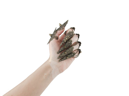 A hand is dramatically adorned with a full set of Myril Jewels studded nail claw rings in bronze. The claw rings feature elaborate filigree with pointed studs, invoking a bold neo-goth aesthetic. These edgy, dark-avantgarde pieces are perfect for those who embrace gothic and alternative styles, making them an ideal choice for Halloween, punk-inspired outfits, or as a statement addition to a whimsigoth or witchcore look. They also serve as eye-catching accessories for minimal goth everyday wear