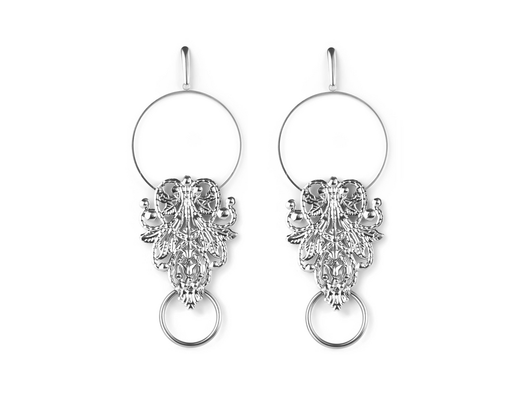 These Myril Jewels earrings feature silver filigree hoops with a baroque flair, blending gothic elegance and punk audacity. The intricate silver filigree contrasts starkly with the bold black detail, culminating in a dangling silver ring, embodying a dark avant-garde aesthetic. Perfect for a neo-gothic look, they are ideal for anyone who loves Halloween, Witchcore, or statement jewelry for festive occasions.