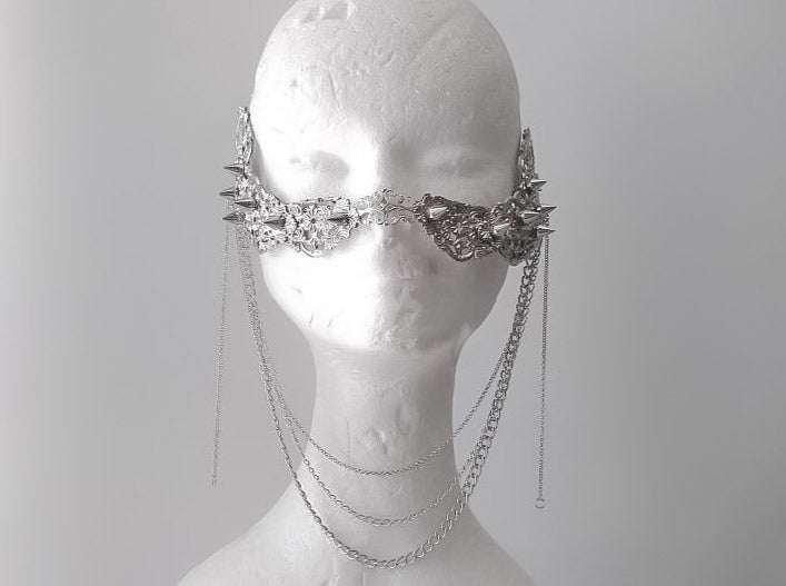 An artistic still life of Myril Jewels' studded nose mask, draped with delicate hanging chains. This piece showcases meticulous craftsmanship, featuring an ornate silver filigree design with spiked details, epitomizing the neo-gothic style. It’s an ideal accessory for those who adore gothic-chic and witchcore fashion, adding an avant-garde edge to Halloween costumes, everyday wear, or as a dramatic accent for rave parties and festival outfits.