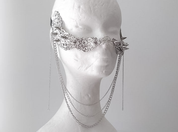 An artistic still life of Myril Jewels' studded nose mask, draped with delicate hanging chains. This piece showcases meticulous craftsmanship, featuring an ornate silver filigree design with spiked details, epitomizing the neo-gothic style. It’s an ideal accessory for those who adore gothic-chic and witchcore fashion, adding an avant-garde edge to Halloween costumes, everyday wear, or as a dramatic accent for rave parties and festival outfits.