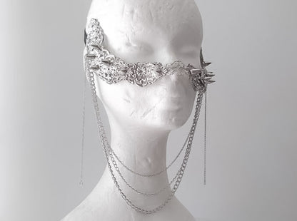 An artistic still life of Myril Jewels' studded nose mask, draped with delicate hanging chains. This piece showcases meticulous craftsmanship, featuring an ornate silver filigree design with spiked details, epitomizing the neo-gothic style. It’s an ideal accessory for those who adore gothic-chic and witchcore fashion, adding an avant-garde edge to Halloween costumes, everyday wear, or as a dramatic accent for rave parties and festival outfits.