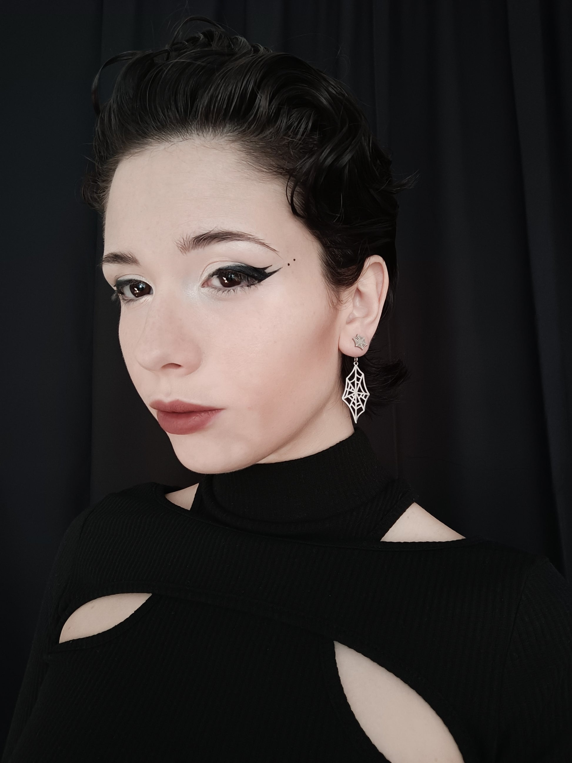 A model exudes gothic elegance with Myril Jewels' spiderweb earrings, a quintessential piece for lovers of neo-goth jewelry. These finely crafted earrings, with their intricate web-like design, are a testament to dark-avantgarde artistry, perfect for adding a touch of gothic-chic to any ensemble. The earrings are showcased against a sleek black outfit, making them ideal for everyday wear, Halloween adornment, or as a distinctive accessory for rave parties and festivals