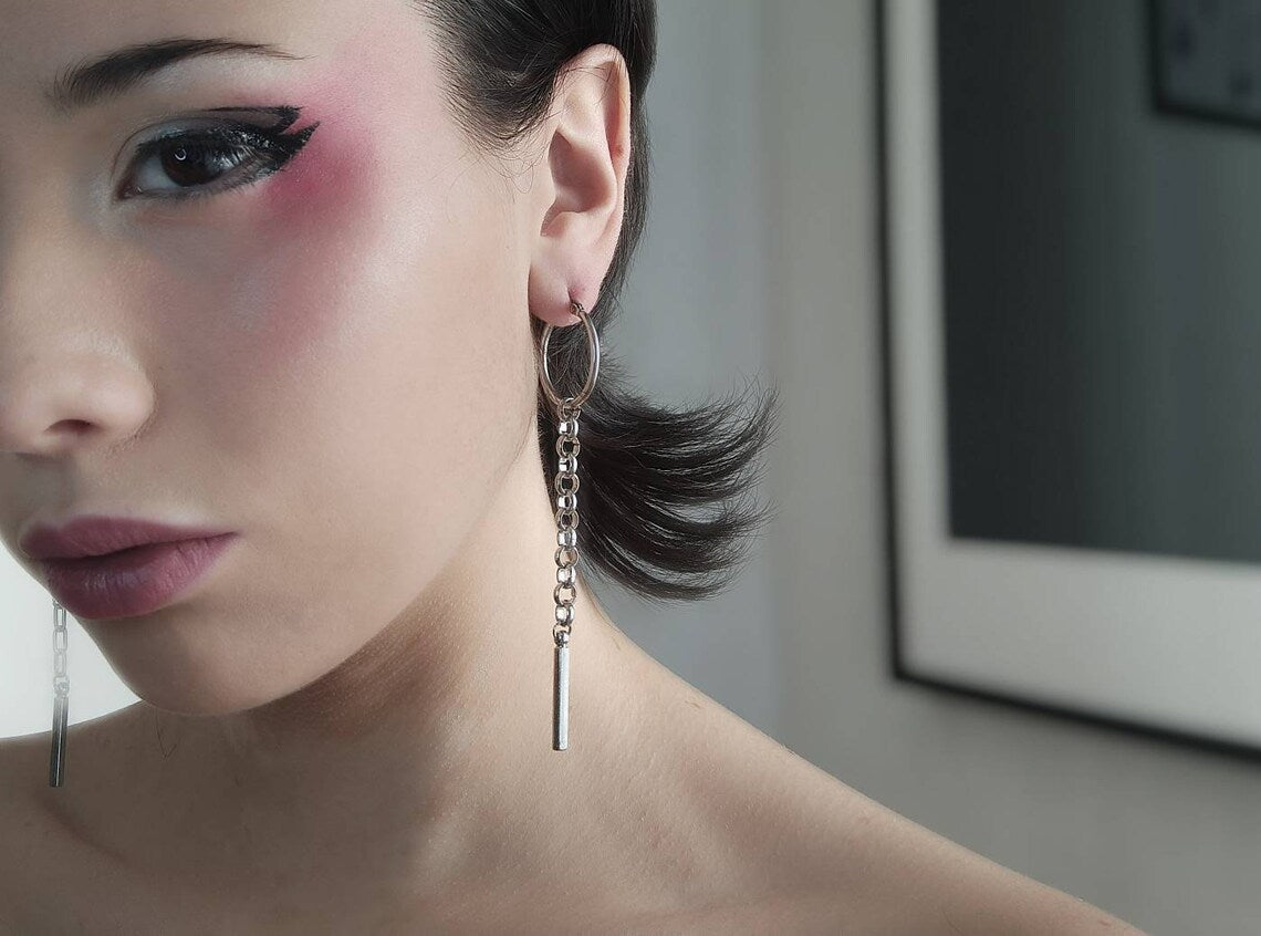 The image presents an elegant model showcasing a pair of Myril Jewels hoop earrings with sleek, long chains. The minimalist design aligns with a dark avant-garde aesthetic, ideal for those who love neo-gothic style, making them a versatile accessory for daily wear or special events like festivals or rave parties. These earrings could also be a bold gift for a goth girlfriend or friend.