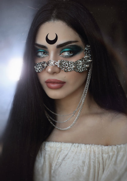 A mysterious allure is captured in this image featuring a model adorned with a Myril Jewels studded nose mask, complete with hanging chains. The mask's elaborate craftsmanship and neo-goth design reflect a dark-avantgarde vibe, perfect for gothic and alternative style aficionados. This unique accessory is a quintessential statement piece for Halloween, rave parties, and festival fashion.