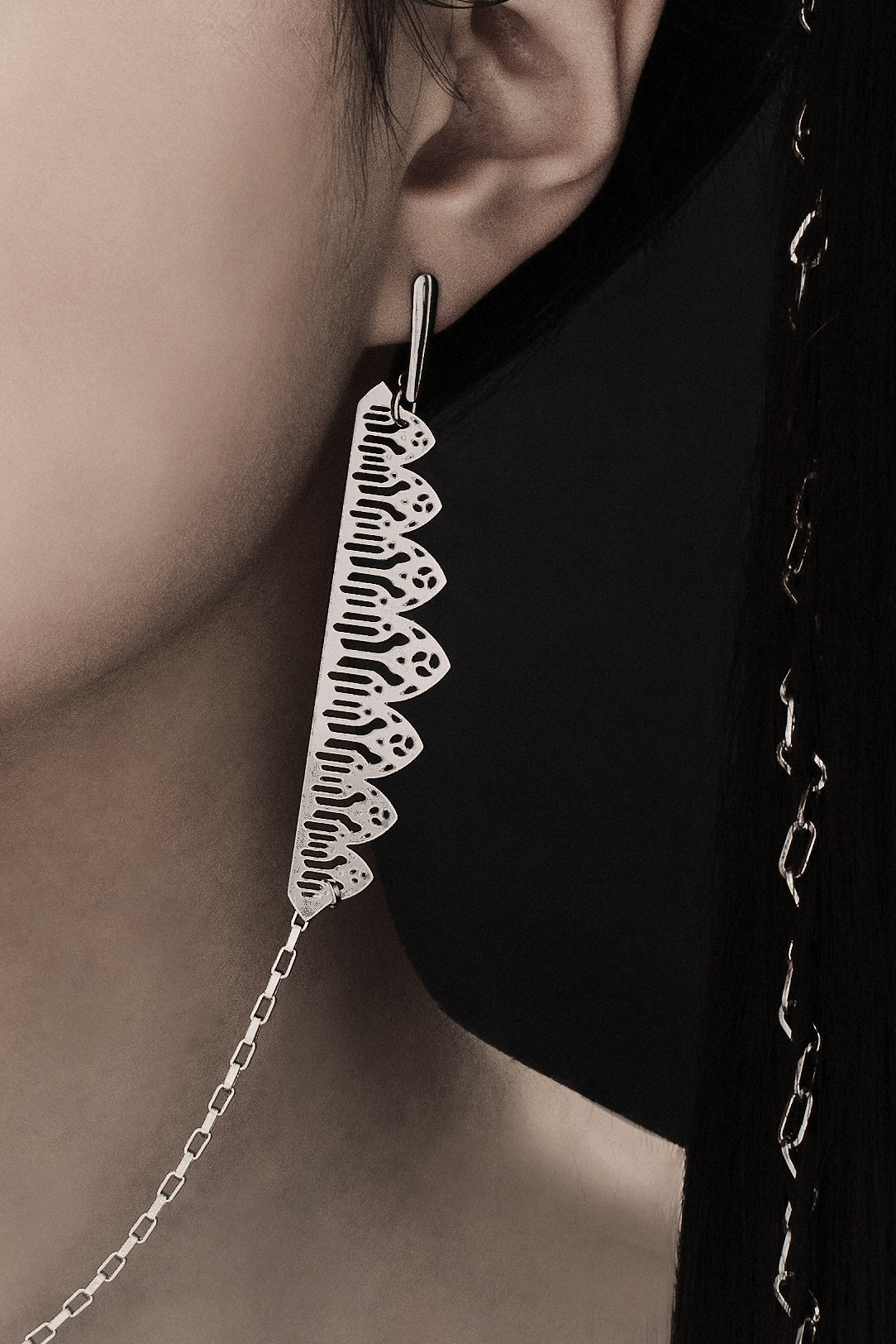 A close up detail of this distinctive single-piece Myril Jewels earring, with an intricate silver design, spans from one ear to the other with a delicate connecting chain. It embodies the essence of neo-gothic elegance, making it an ideal accessory for minimal goth everyday wear, or as a standout piece for rave parties. This earring captures the gothic-chic trend and is a unique gift idea for those with a love for bold, alternative jewelry
