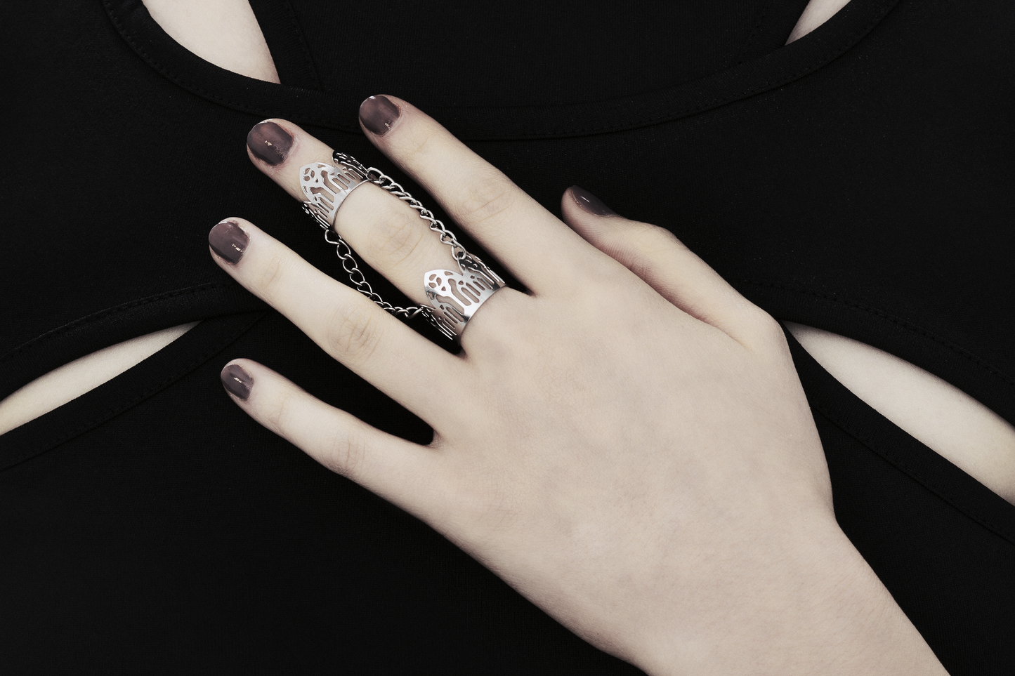 A model's hand is adorned with a Myril Jewels double ring, inspired by gothic arches, against a black top with cut-out details. The ring showcases the brand's dark-avantgarde style, ideal for gothic and alternative fashion aficionados. With a design perfect for Halloween, the ring also suits everyday wear, contributing to a minimal goth look or as an accent for rave party and festival attire. It embodies the gothic-chic and whimsigoth essence, making it a unique addition to any witchcore jewelry collection