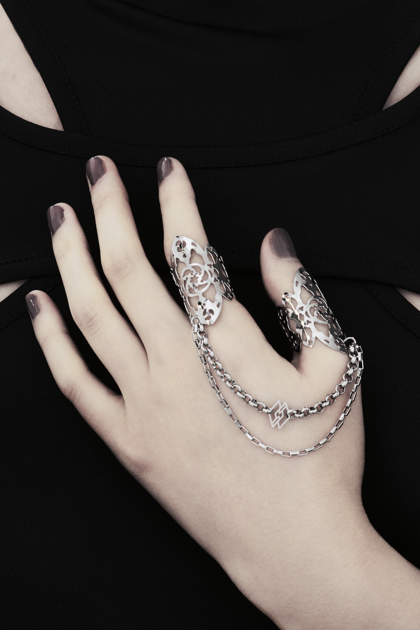 A hand models Myril Jewels' handcrafted ring featuring elaborate cut-out designs that echo a gothic architectural aesthetic, suited for lovers of dark-avantgarde fashion. The rings are interconnected with sleek chains, creating a bold, edgy look. This collection exemplifies the Italian craftsmanship and innovative style synonymous with the Myril Jewels brand, catering to the tastes of the alternative and gothic jewelry community.