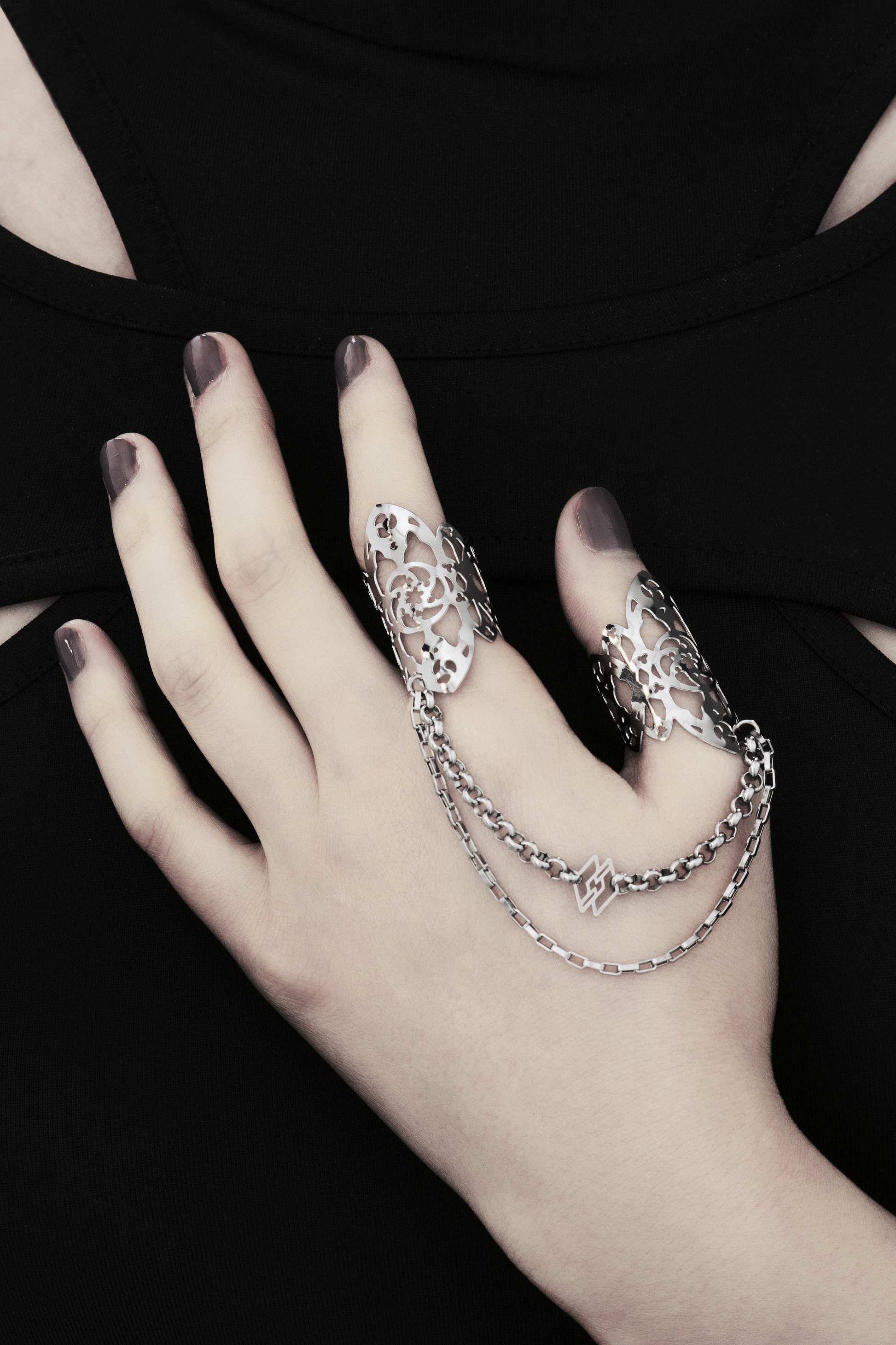 A hand models Myril Jewels' handcrafted ring featuring elaborate cut-out designs that echo a gothic architectural aesthetic, suited for lovers of dark-avantgarde fashion. The rings are interconnected with sleek chains, creating a bold, edgy look. This collection exemplifies the Italian craftsmanship and innovative style synonymous with the Myril Jewels brand, catering to the tastes of the alternative and gothic jewelry community.