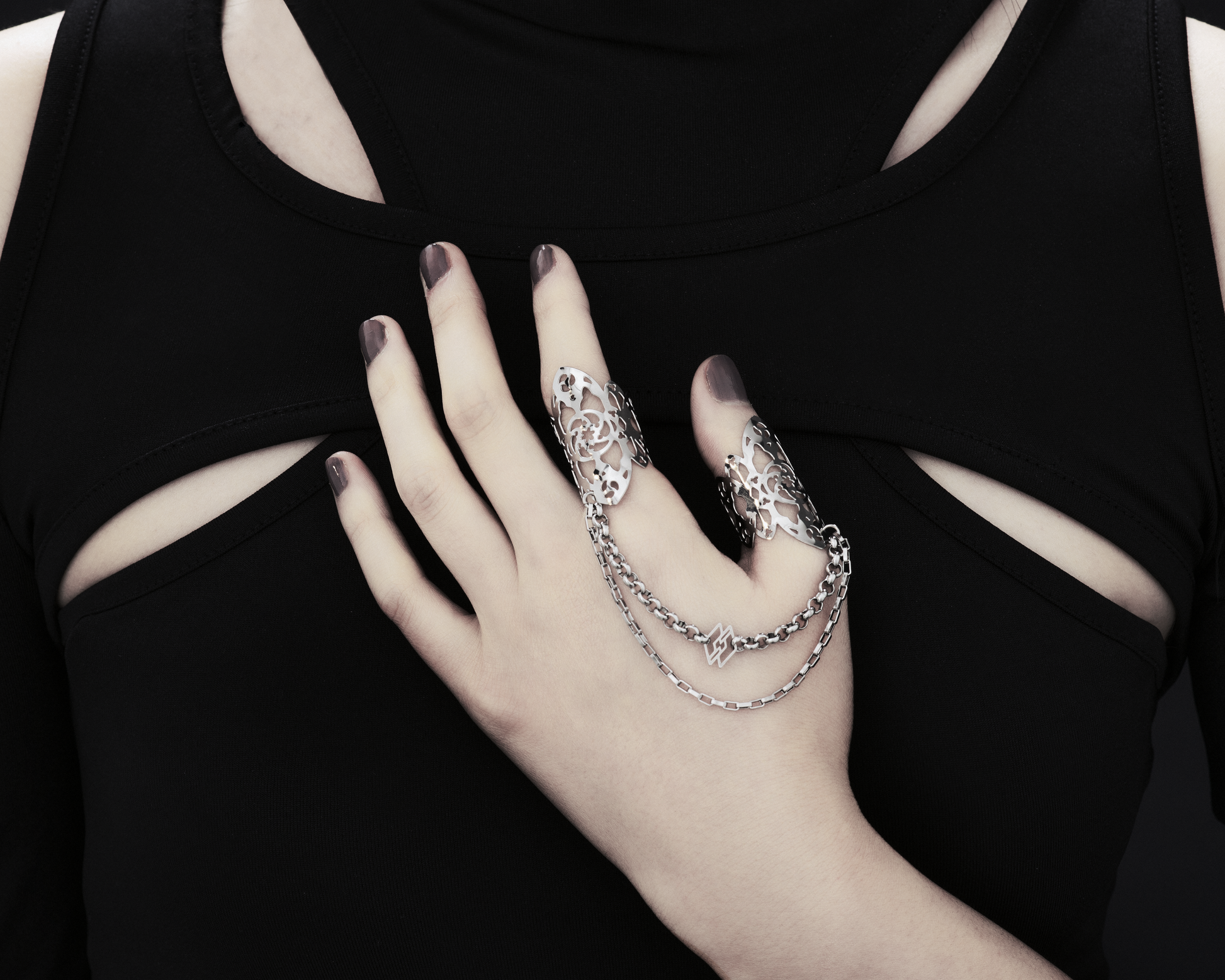 A hand models Myril Jewels' handcrafted ring featuring elaborate cut-out designs that echo a gothic architectural aesthetic, suited for lovers of dark-avantgarde fashion. The rings are interconnected with sleek chains, creating a bold, edgy look. This collection exemplifies the Italian craftsmanship and innovative style synonymous with the Myril Jewels brand, catering to the tastes of the alternative and gothic jewelry community.