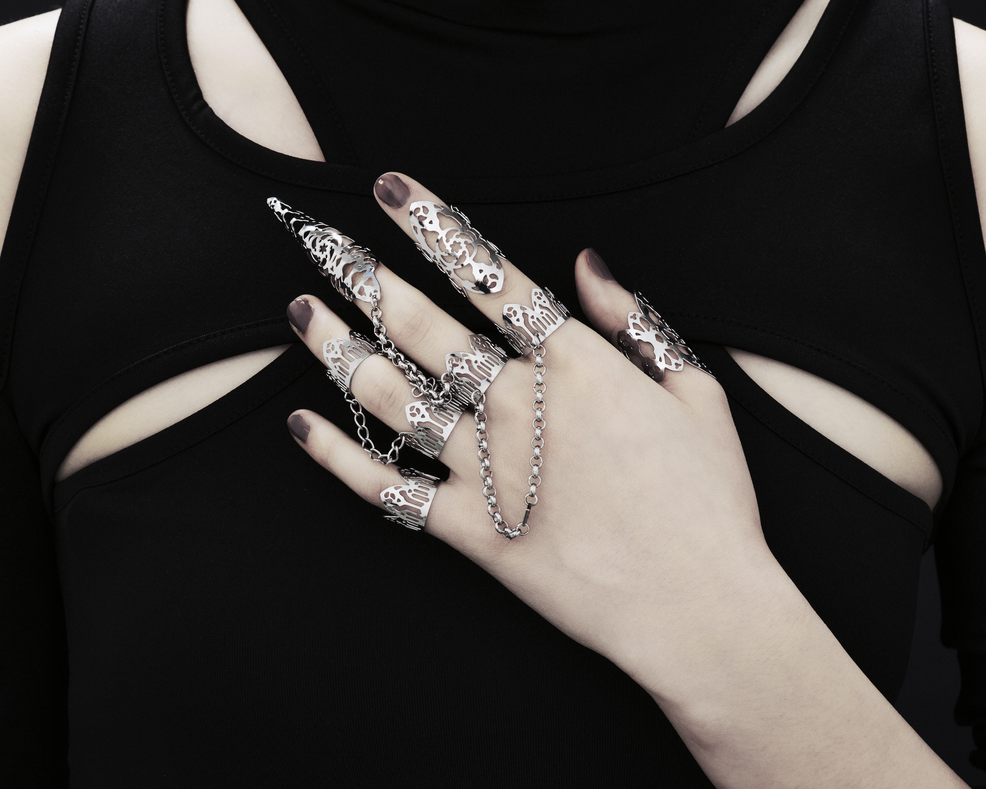 A hand models an array of Myril Jewels' handcrafted rings, each piece featuring elaborate cut-out designs that echo a gothic architectural aesthetic, suited for lovers of dark-avantgarde fashion. The rings, varying in size from slender bands to elongated claw-like shapes, are interconnected with sleek chains, creating a bold, edgy look. This collection exemplifies the Italian craftsmanship and innovative style synonymous with the Myril Jewels brand, catering to the tastes of the gothic community.