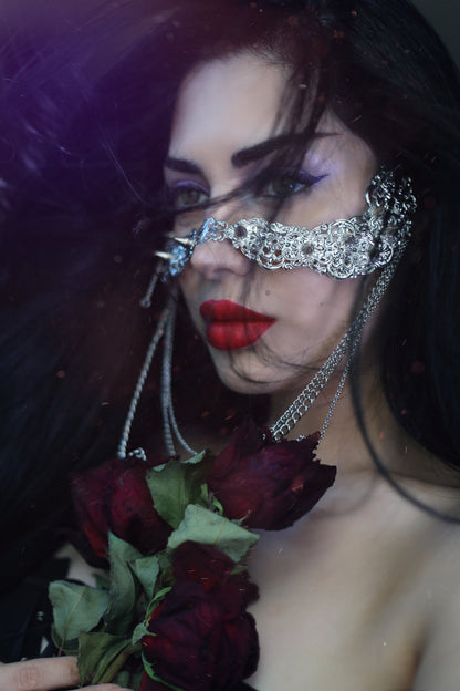 A model from Myril Jewels exudes dark-avantgarde sophistication while wearing a studded nose mask adorned with hanging chains. The mask, paired with the model's bold red lips and a bouquet of deep red roses, creates a captivating and mysterious look, perfect for Halloween, everyday wear, or as an exceptional accessory for rave parties and festivals.