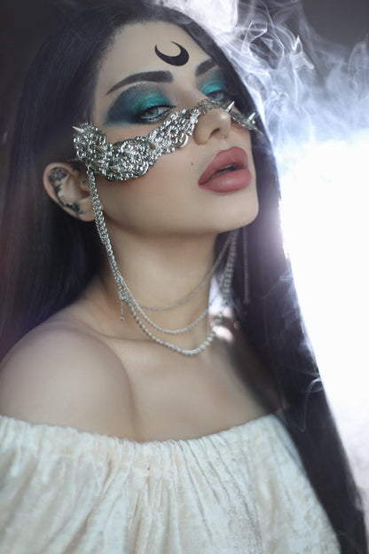 In this striking portrait, a model presents a Myril Jewels studded nose mask adorned with hanging chains, embodying the brand's neo-goth aesthetic. The mask, a fusion of punk and gothic-chic elements, is a daring piece for those who embrace dark-avantgarde fashion. It's a statement accessory, perfect for Halloween, witchcore styles, or as a distinctive highlight to minimalist goth attire, showcasing Myril's dedication to unique, alternative jewelry designs